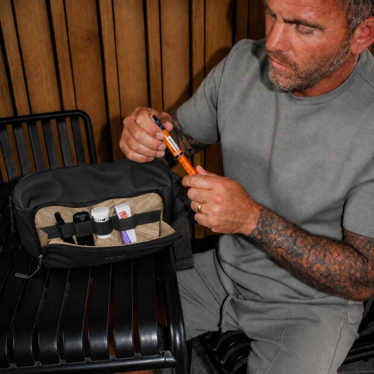 A man in a grey t-shirt sits on a black metal bench, holding an insulin pen next to an open black and beige travel pouch containing small medical or utility items.