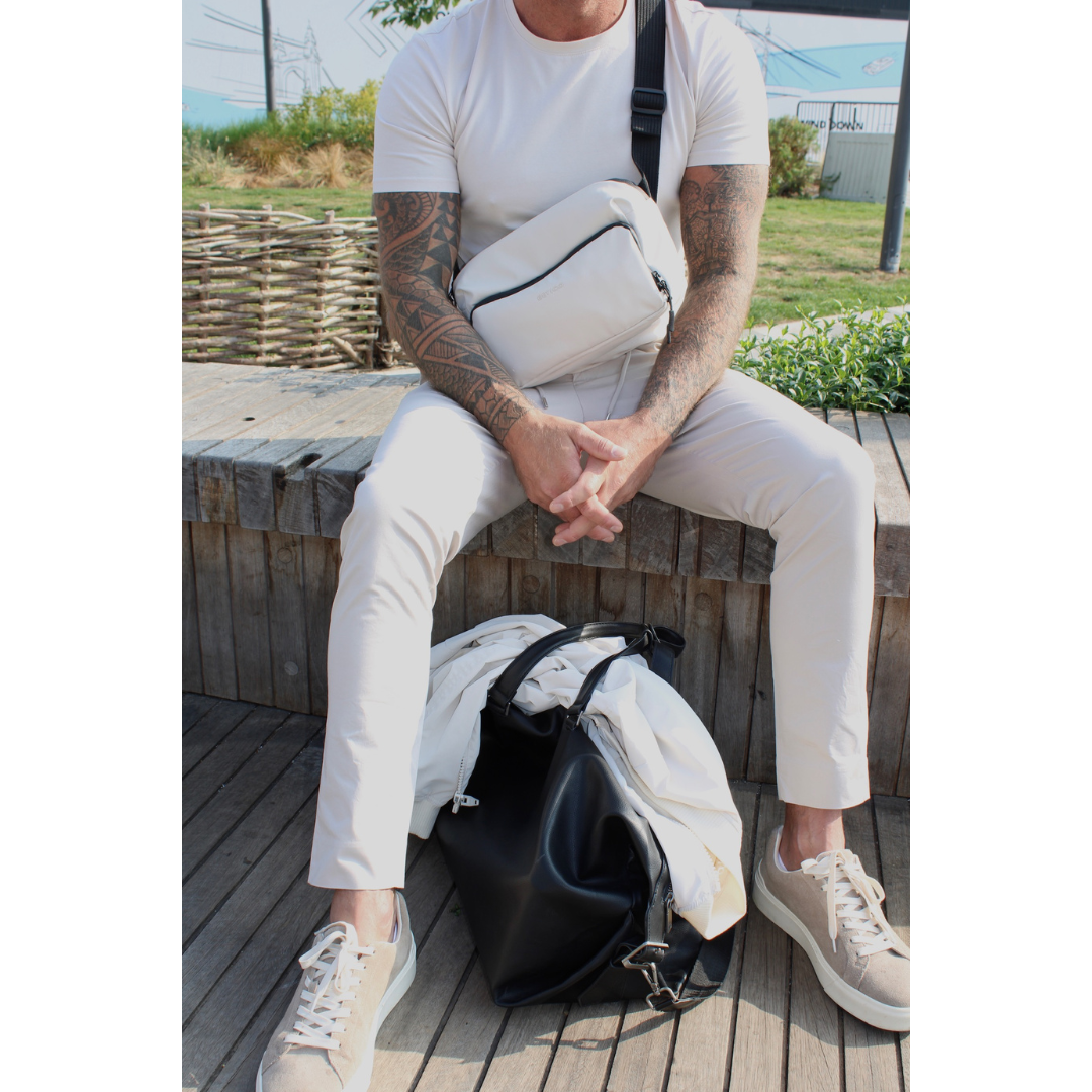 Man sitting outdoors wearing the Betico Essential Bag in cream as a discreet crossbody bag with the black Betico Essential Bag beside him for everyday travel and organised daily carry