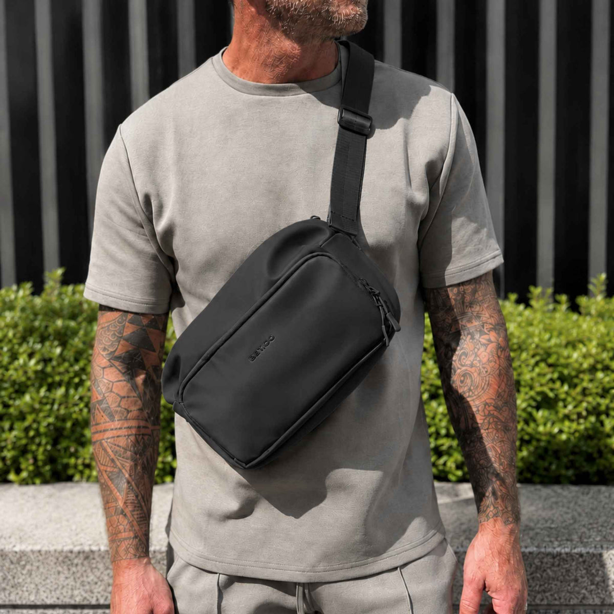 A man wearing a black crossbody sling bag with an adjustable shoulder strap, standing outdoors in front of a dark slatted wall and greenery.