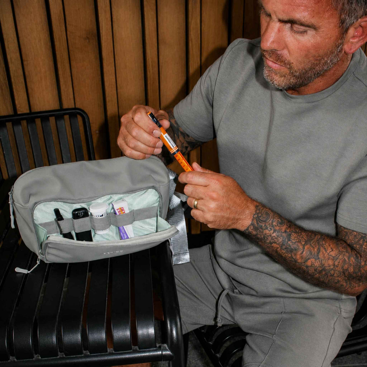 Man seated beside an open green Betico Essential Diabetes Bag, handling an insulin pen with organised diabetes supplies visible in the front compartment.